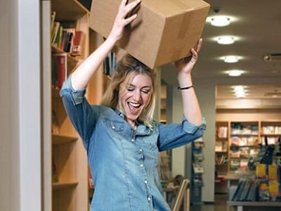 A woman in a bookstore joyfully holds a large package in the air