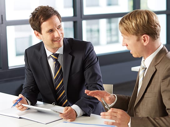 Two men in suits sit next to each other at the table and talk business