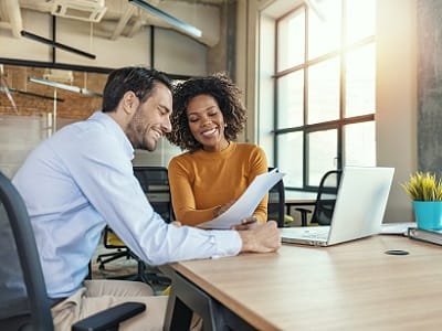 A man and a woman are sitting in an office. She shows him documents and has the laptop open next to her