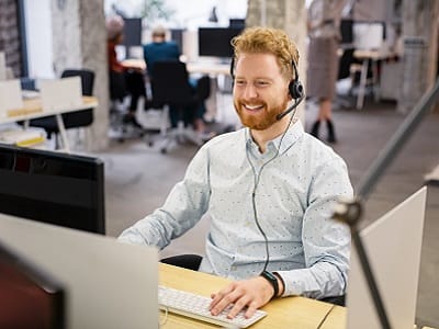 A man in a shirt is happily talking on the headset to a customer while sitting at the PC