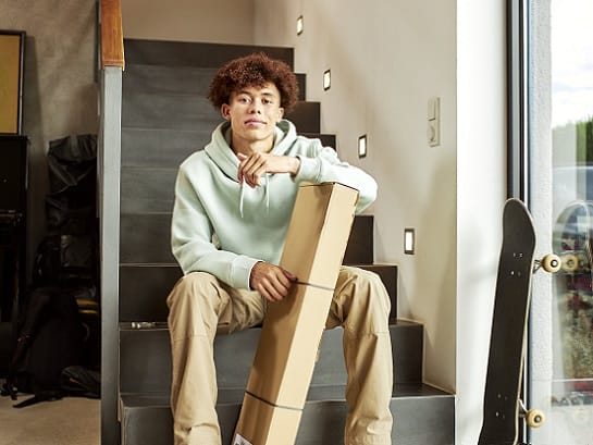 Ein Teenager sitzt auf der Treppe im Haus und stÃ¼tz sich auf ein langes Paket ab. Neben ihm steht ein neues Skateboard