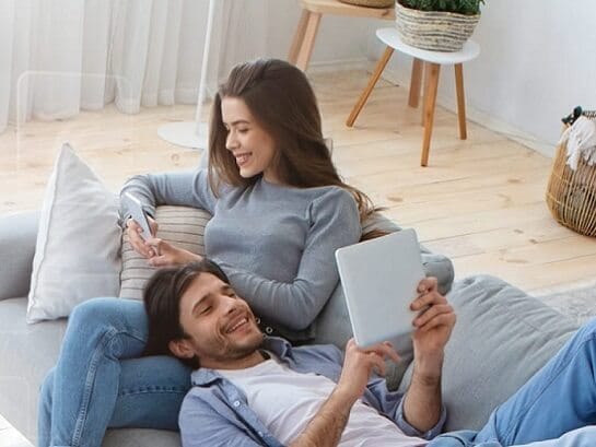 A woman sits on the couch and looks at the delivery options of GLS on her smartphone. On her lap is her husband who is at the tablet