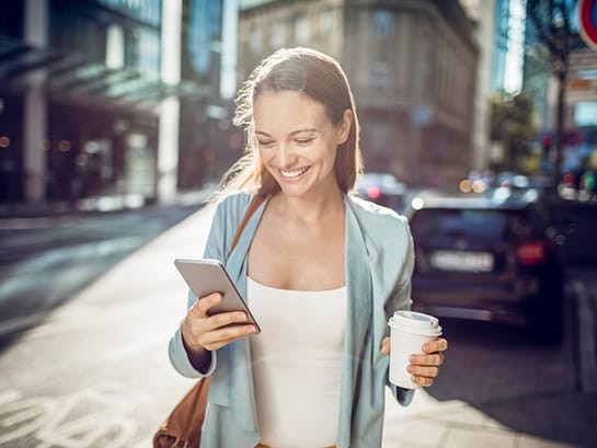 A woman holds a coffee to go in one hand and uses her smartphone to change the delivery option of her package with the other.