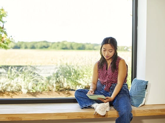 Eine junge Frau mit dunklen langen Haaren sitz auf der Fensterbank und surft mit ihrem Tablet auf der GLS Paket Seite